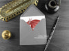 Welsh dragon greeting card, shown lying flat, with my original pen and ink illustration of a red dragon, inside an open grey envelope. With vintage Victorian pen and writing desk accessories. Design and illustration © Nicola L Robinson | Teeth and Claws