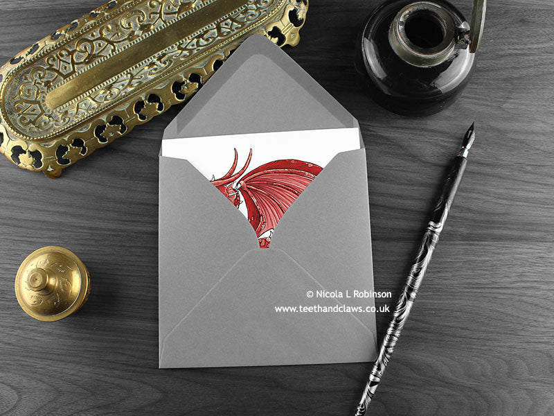 Welsh dragon greeting card, shown lying flat, with my original pen and ink illustration of a red dragon, inside an open grey envelope. With vintage Victorian pen and writing desk accessories. Design and illustration © Nicola L Robinson | Teeth and Claws