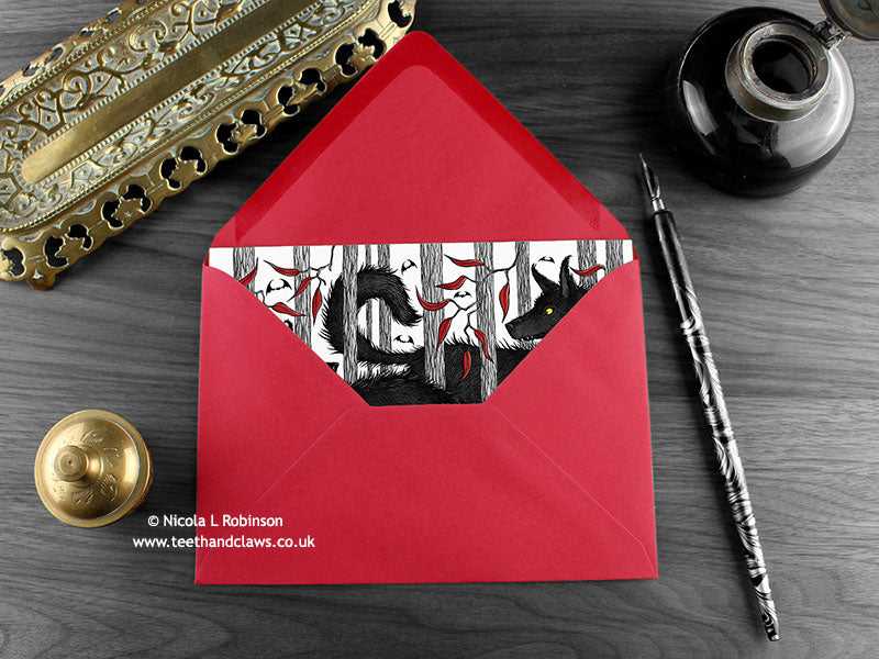 Wolves love card, shown lying flat, with my original pen and ink illustration of two wolves, inside an open red envelope. With vintage Victorian pen and writing desk accessories. Design and illustration © Nicola L Robinson | Teeth and Claws