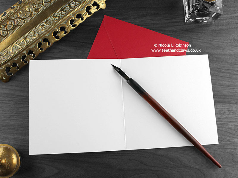 Ankylosaurus Christmas card shown open and flat with blank inside, on a red envelope, alongside vintage Victorian pen and writing desk accessories. Design and illustration © Nicola L Robinson | Teeth and Claws
