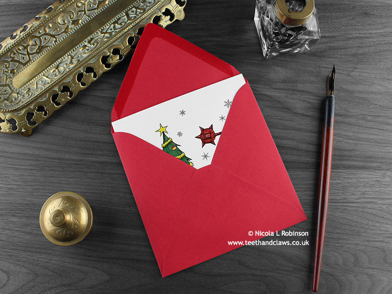 Dinosaur Christmas card, shown lying flat, with my original pen and ink illustration of a festive ankylosaurus, inside an open red envelope. With vintage Victorian pen and writing desk accessories. Design and illustration © Nicola L Robinson | Teeth and Claws