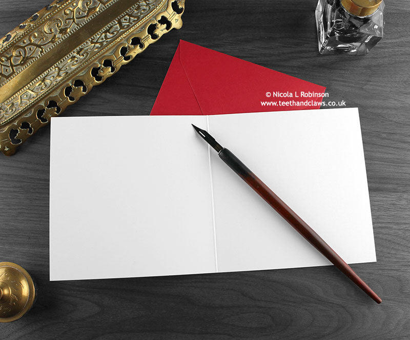 Dinosaur Christmas card, shown open and flat with blank inside, on a red envelope, alongside vintage Victorian pen and writing desk accessories. Design and illustration © Nicola L Robinson | Teeth and Claws