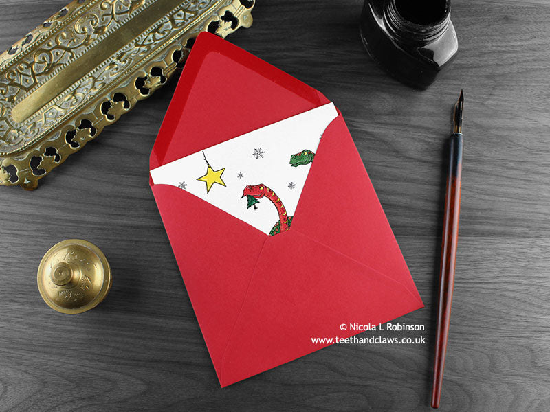 Dinosaur Christmas card, shown lying flat, with my original pen and ink illustration of festive diplodocus, inside an open red envelope. With vintage Victorian pen and writing desk accessories. Design and illustration © Nicola L Robinson | Teeth and Claws