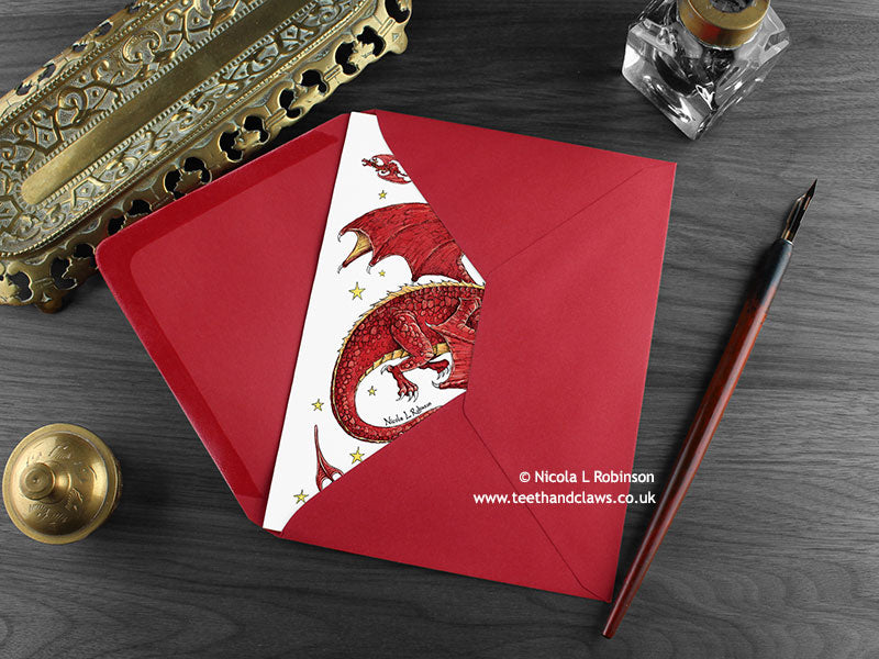 Dragon Christmas card, shown lying flat, with my original pen and ink illustration of a festive red dragon, inside an open red envelope. With vintage Victorian pen and writing desk accessories. Design and illustration © Nicola L Robinson | Teeth and Claws