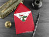 Dragon Christmas card, shown lying flat, with my original pen and ink illustration of a festive green dragon, inside an open red envelope. With vintage Victorian pen and writing desk accessories. Design and illustration © Nicola L Robinson | Teeth and Claws