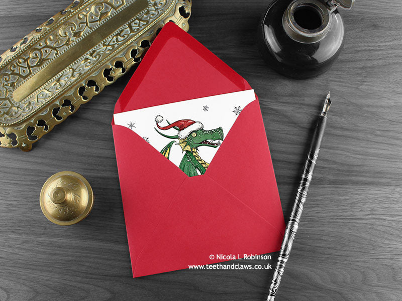 Dragon Christmas card, shown lying flat, with my original pen and ink illustration of a festive green dragon, inside an open red envelope. With vintage Victorian pen and writing desk accessories. Design and illustration © Nicola L Robinson | Teeth and Claws