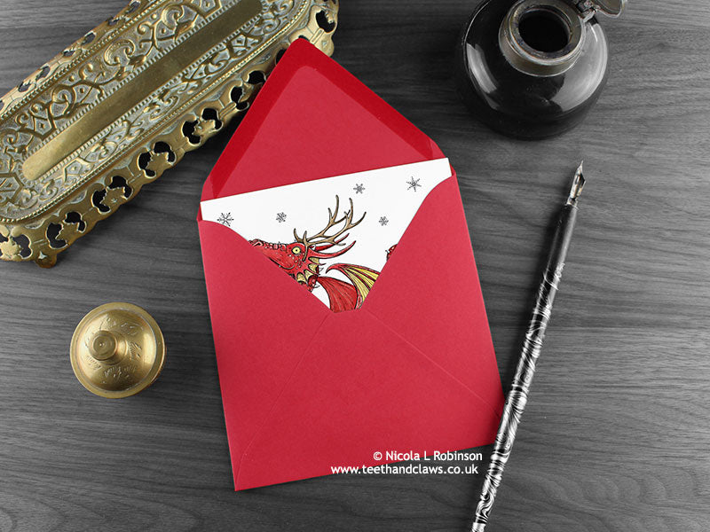 Dragon Christmas card, shown lying flat, with my original pen and ink illustration of a festive red dragon, inside an open red envelope. With vintage Victorian pen and writing desk accessories. Design and illustration © Nicola L Robinson | Teeth and Claws