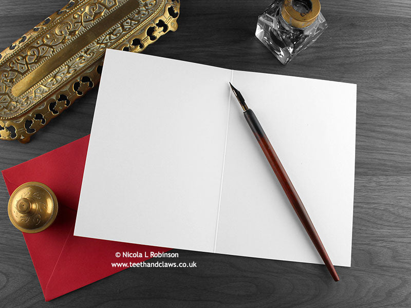Dragon Christmas card, shown open and flat with blank inside, on a red envelope, alongside vintage Victorian pen and writing desk accessories. Design and illustration © Nicola L Robinson | Teeth and Claws