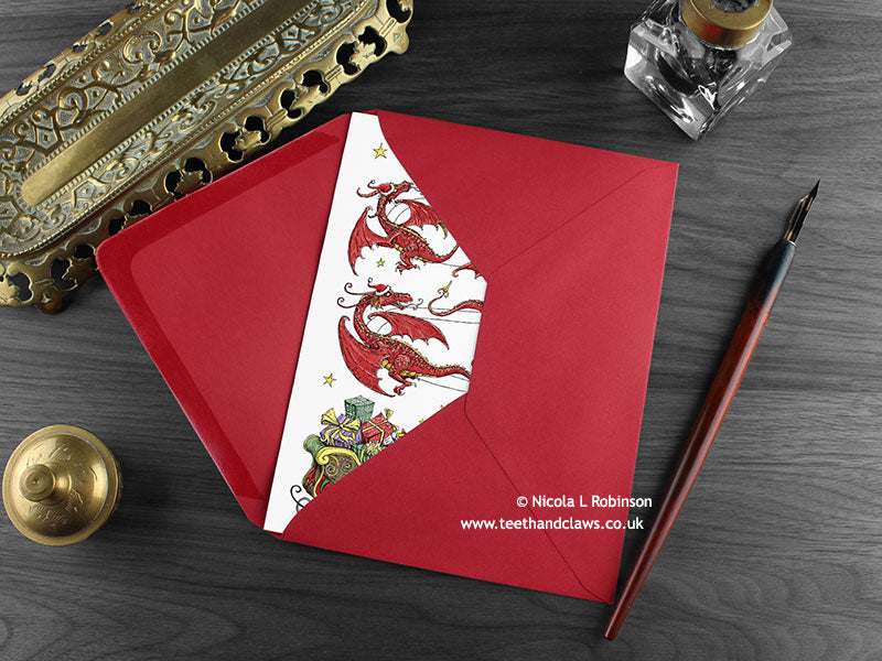 Dragon Christmas card, shown lying flat, with my original pen and ink illustration of festive dragons, inside an open red envelope. With vintage Victorian pen and writing desk accessories. Design and illustration © Nicola L Robinson | Teeth and Claws