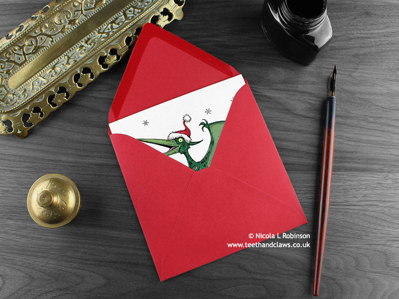 Pteranodon Christmas card, shown lying flat, with my original pen and ink illustration of a festive pteranodon, inside an open red envelope. With vintage Victorian pen and writing desk accessories. Design and illustration © Nicola L Robinson | Teeth and Claws