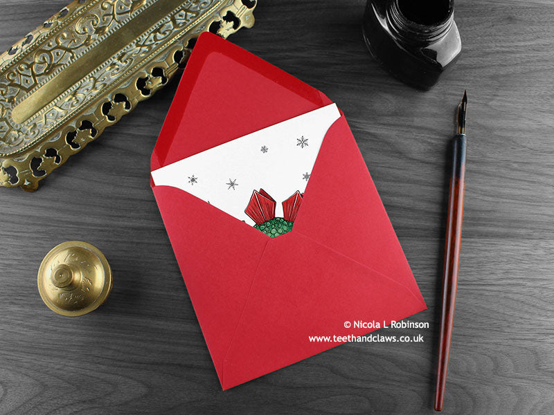 Stegosaurus Christmas card, shown lying flat, with my original pen and ink illustration of a festive stegosaurus, inside an open red envelope. With vintage Victorian pen and writing desk accessories. Design and illustration © Nicola L Robinson | Teeth and Claws