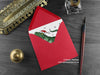 Dinosaur Christmas card, shown lying flat, with my original pen and ink illustration of a festive T rex, inside an open red envelope. With vintage Victorian pen and writing desk accessories. Design and illustration © Nicola L Robinson | Teeth and Claws