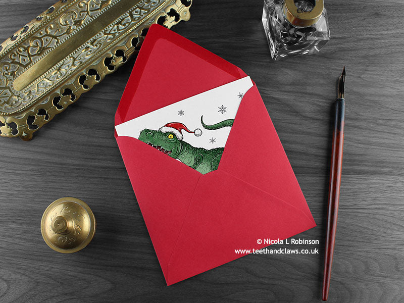 Dinosaur Christmas card, shown lying flat, with my original pen and ink illustration of a festive T rex, inside an open red envelope. With vintage Victorian pen and writing desk accessories. Design and illustration © Nicola L Robinson | Teeth and Claws