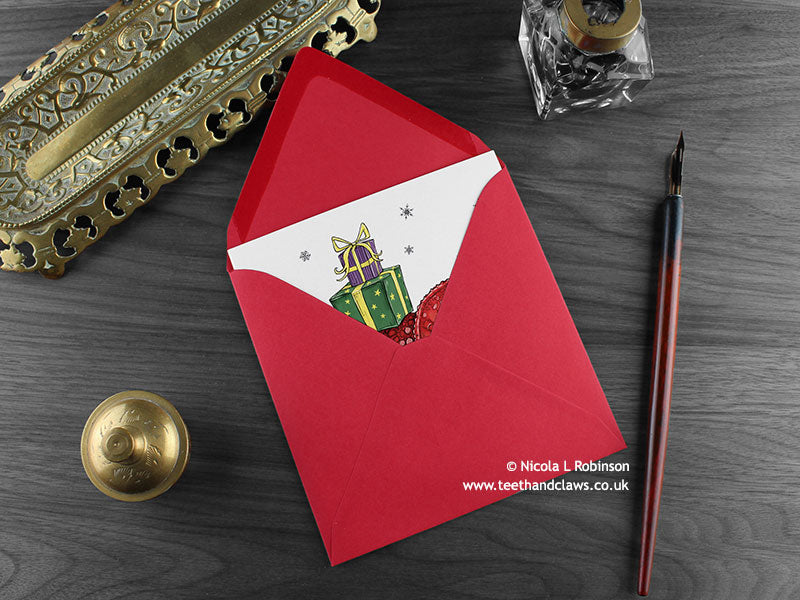 Dinosaur Christmas card, shown lying flat, with my original pen and ink illustration of a festive triceratops, inside an open red envelope. With vintage Victorian pen and writing desk accessories. Design and illustration © Nicola L Robinson | Teeth and Claws